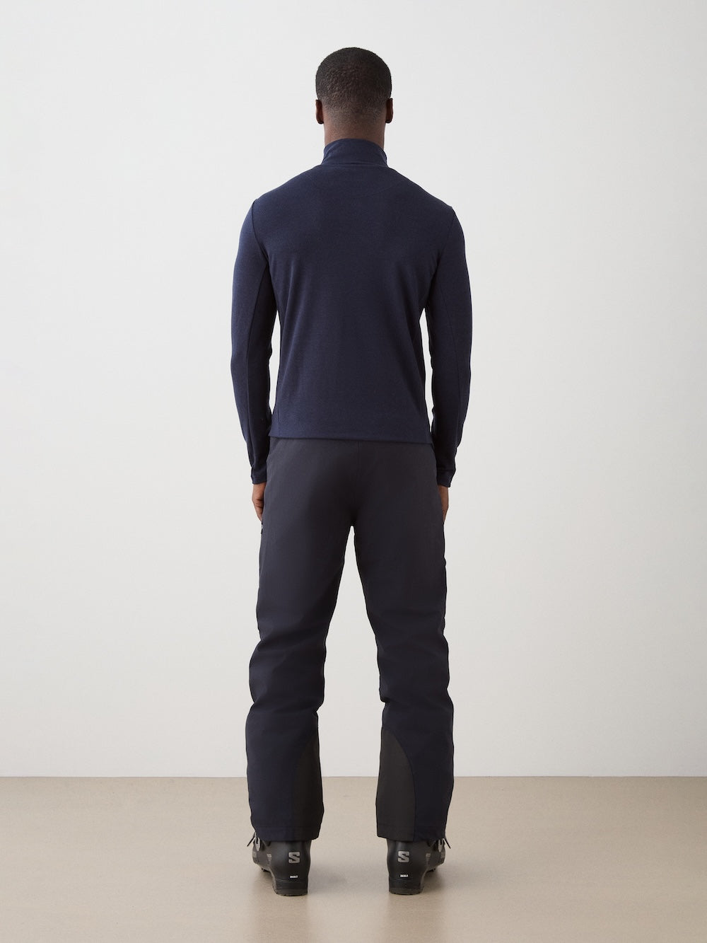 A man faces away from the camera, wearing the We Norwegians Ski 1/4-Zip Men in navy with dark pants and black boots against a plain light background.