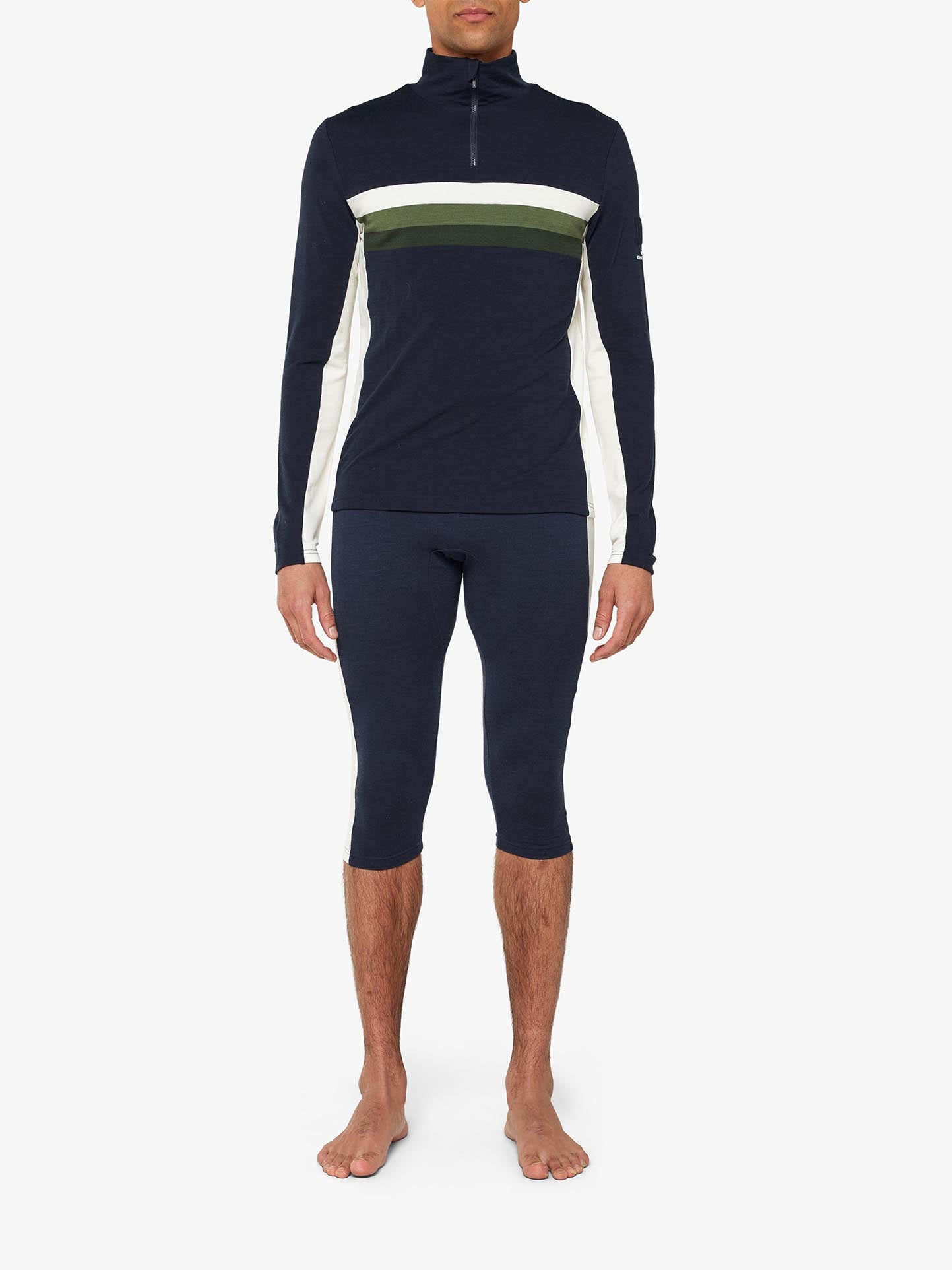 A person is standing barefoot wearing a matching dark navy athletic outfit consisting of the "Ski 1/4-Zip Men" by We Norwegians, a long-sleeved, retro-inspired base layer with horizontal white and olive green stripes across the chest, paired with three-quarter length fitted pants. The background is plain white.