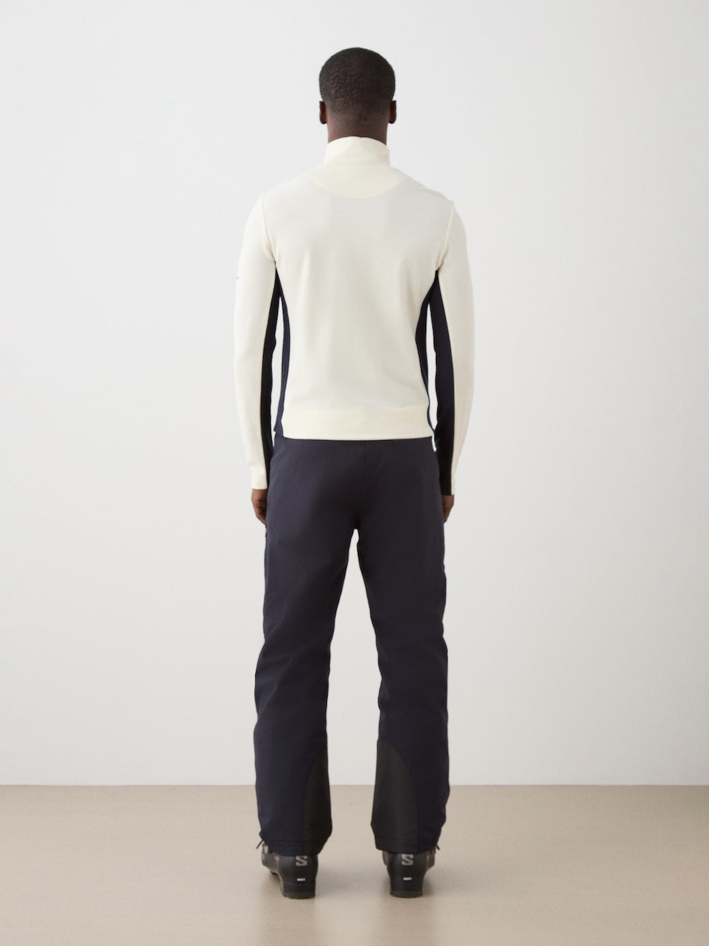 A person stands with their back to the camera, wearing the We Norwegians Ski 1/4-Zip Men merino wool base layer in white, paired with dark pants and black shoes, against a plain light background.