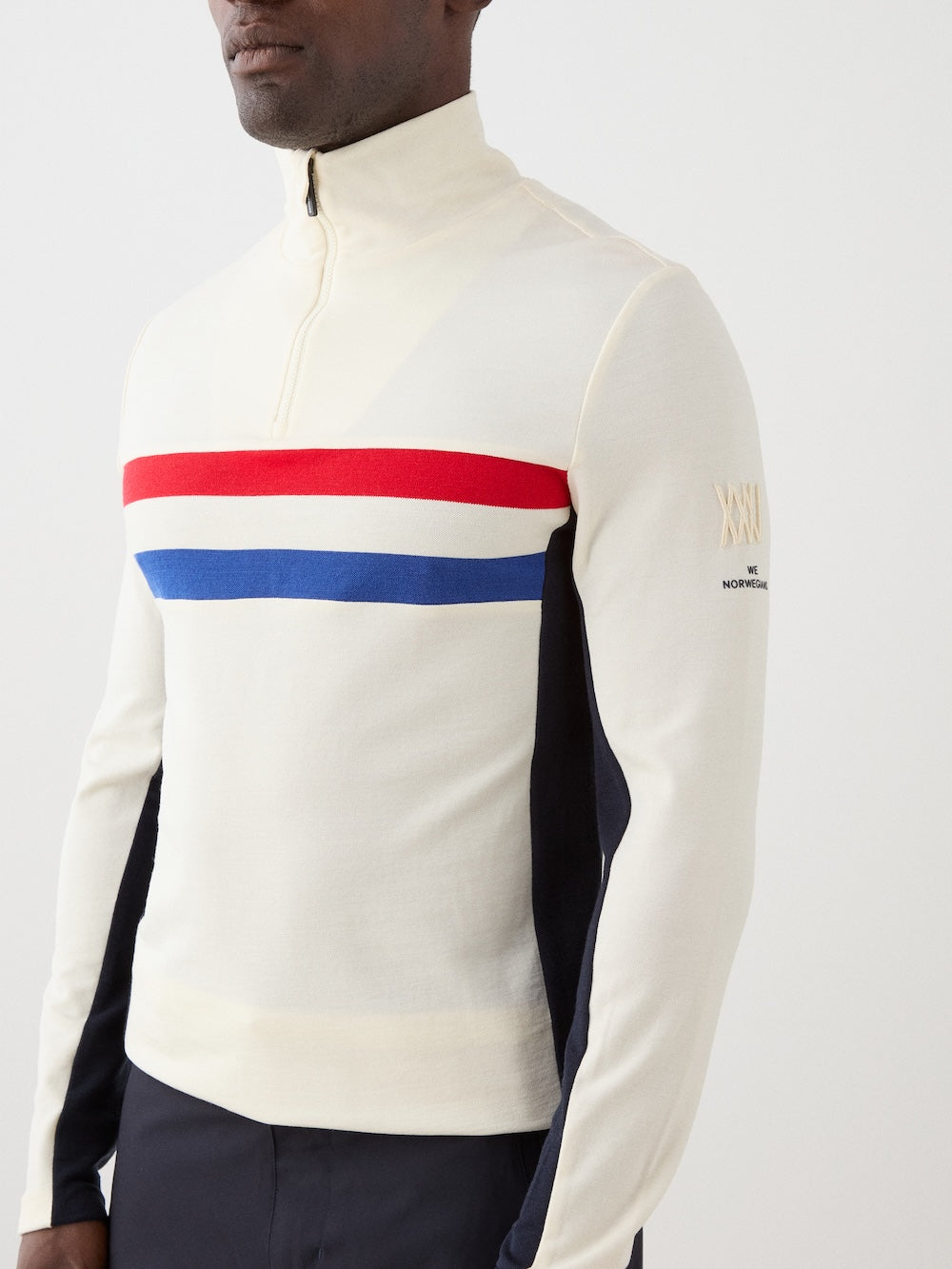 A man wears the We Norwegians Ski 1/4-Zip Men sweater, featuring a cream color, red and blue chest stripes, and "XXIV" embroidered on the left sleeve.