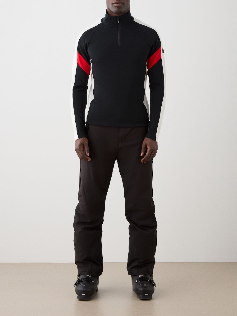 The We Norwegians Zermatt Zipup Men is a Merino wool ski sweater with a half-zip collar, black body, cream sleeves with black inner arms, diagonal red stripes, and a circular patch on the left sleeve | Front