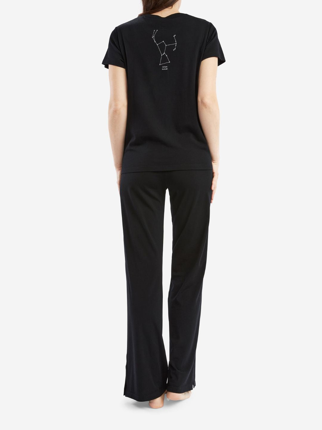 A person with long hair is seen from behind, wearing limited edition We Norwegians Natt PJs Pants Women Black and a matching black short-sleeve top. The back of the top features a simple white line drawing of the Ursa Major constellation. The person is standing barefoot on a plain white background.