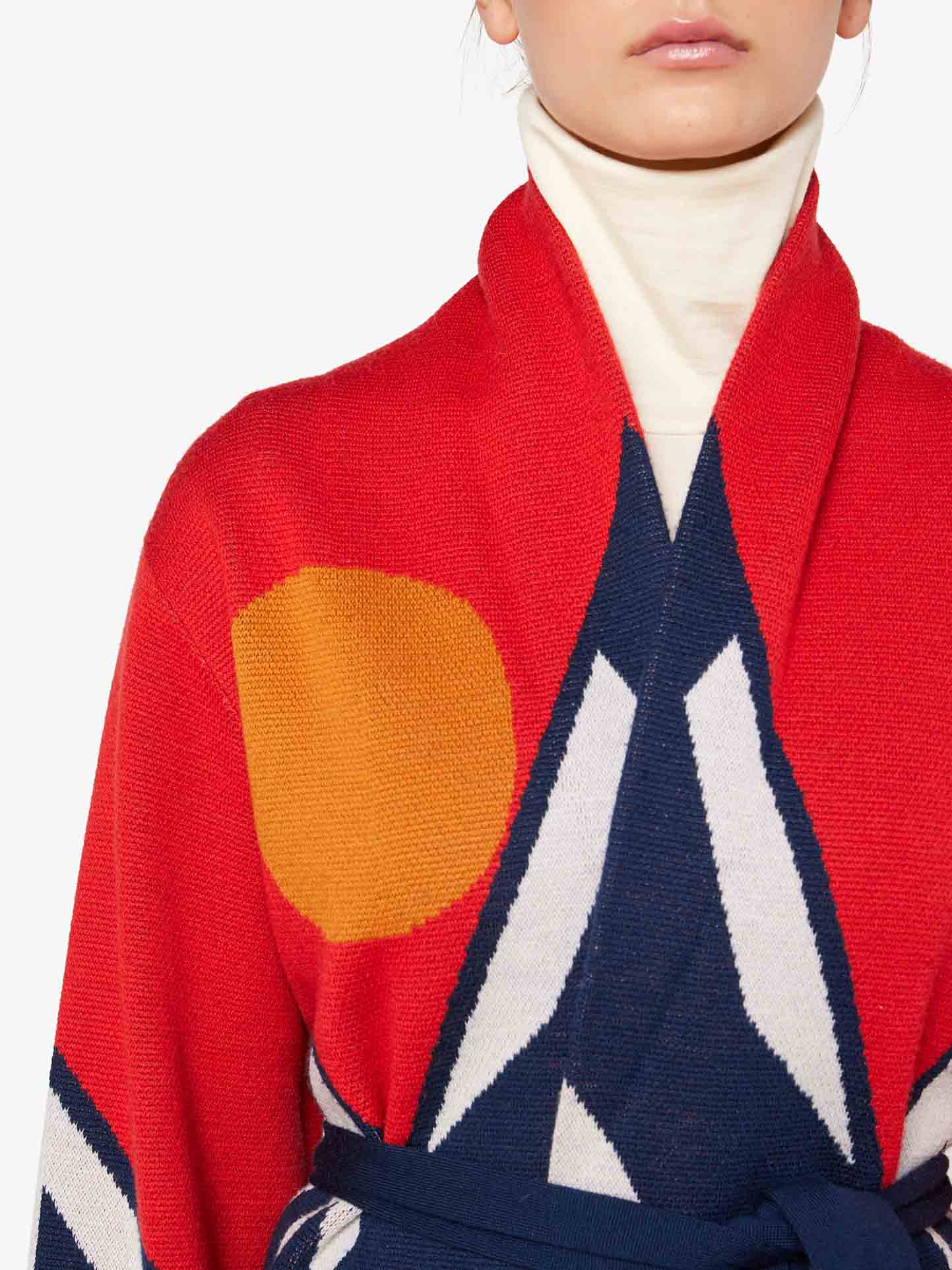 A person is wearing the We Norwegians Snowmountain Cardigan Women, a red sweater with a bold graphic design that features a large yellow circle and navy blue accents, evoking an après-ski style. Underneath, they have on a white turtleneck. The person is visible from the shoulders to just below the nose.