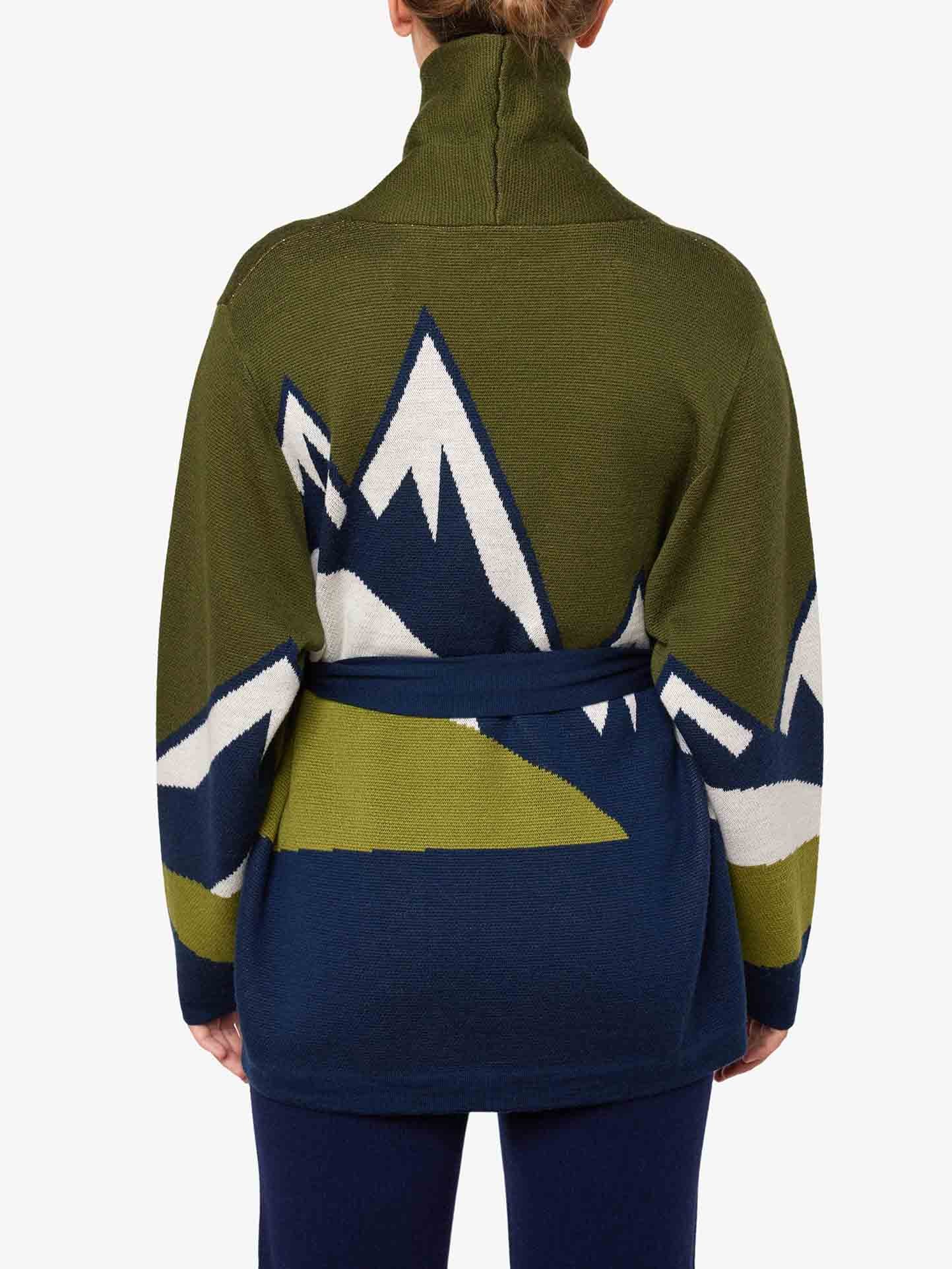 A person is shown from the back wearing the We Norwegians Snowmountain Cardigan Women in green, crafted from merino wool. This high-collared cardigan features long sleeves and a waist belt. Perfect for an aprés ski look, it showcases a striking blue and white mountain pattern on the back. The individual has light brown hair tied up.