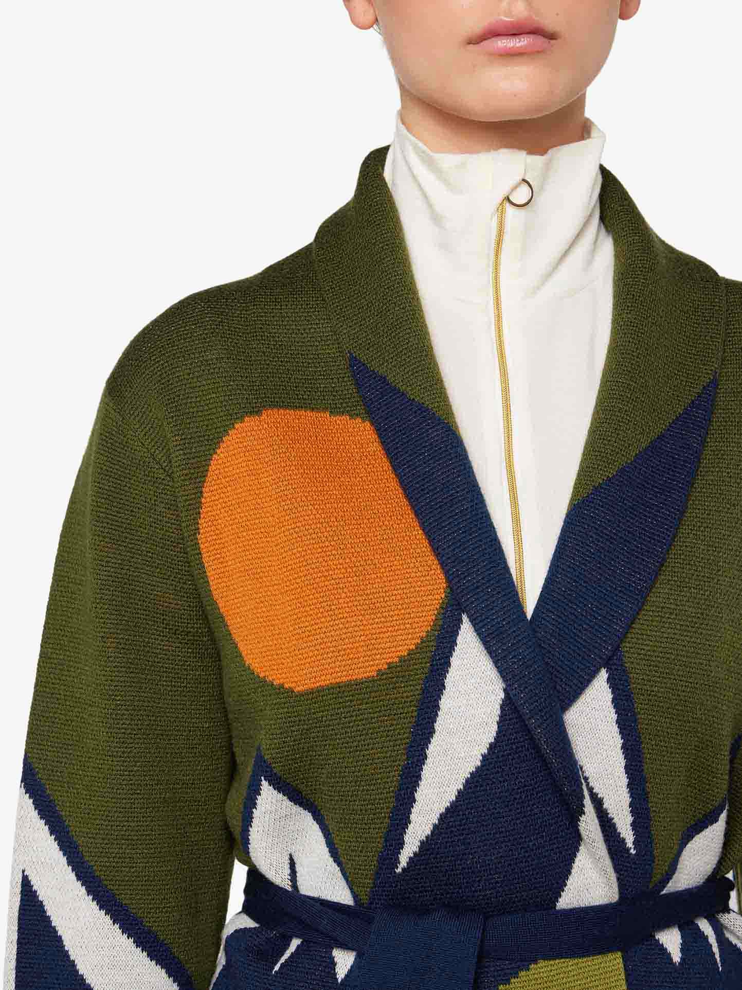 A person is seen wearing the We Norwegians Snowmountain Cardigan Women, which features large abstract patterns in blue, white, and orange on a green background, giving off an après ski look. Underneath the Merino wool cardigan, they have on a high-collared, zip-up white top. The image captures the person's upper torso and part of their head, but the face is not fully visible.