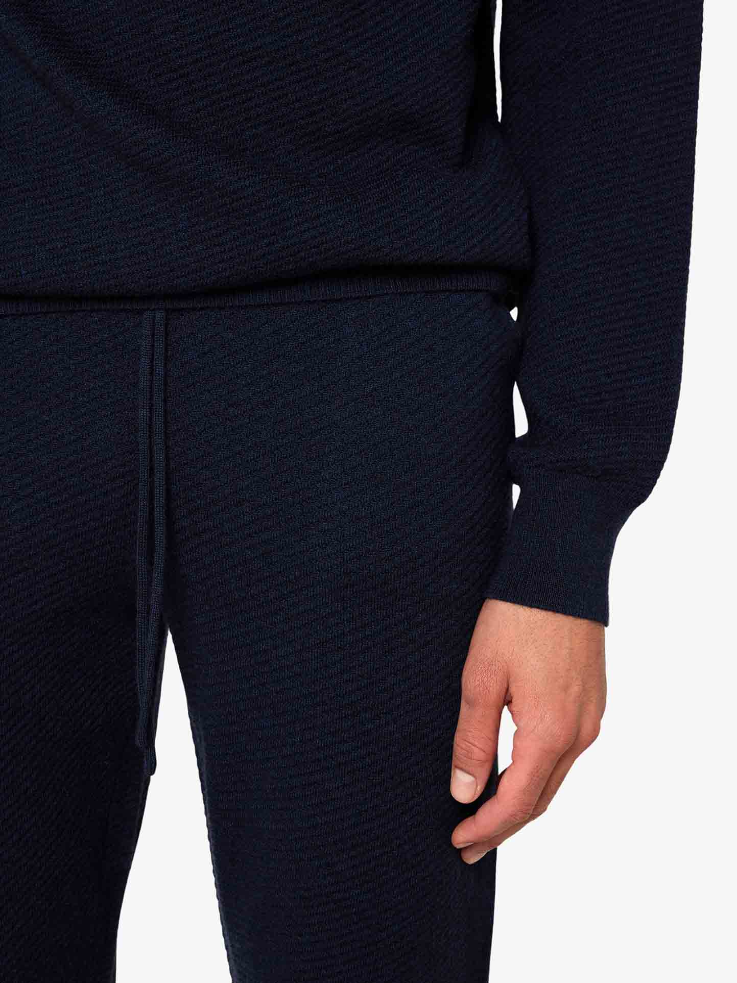 A person is shown wearing the Geilo Jogger Men by We Norwegians, a dark-colored knit lounge set made of fine Merino wool. The image focuses on the lower part of their torso and one hand hanging by their side against a plain white background.