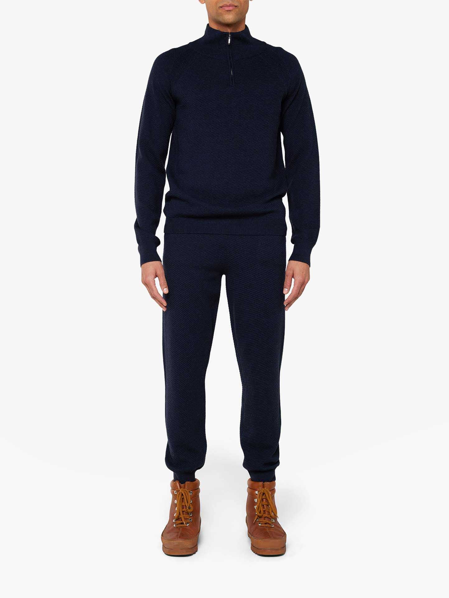 A person wearing the We Norwegians Geilo Jogger Men set in dark blue, featuring ribbed cuffs and an adjustable waist, stands facing the camera. The sweater boasts a half-zip design. They are also sporting light brown boots, all set against a plain white background.