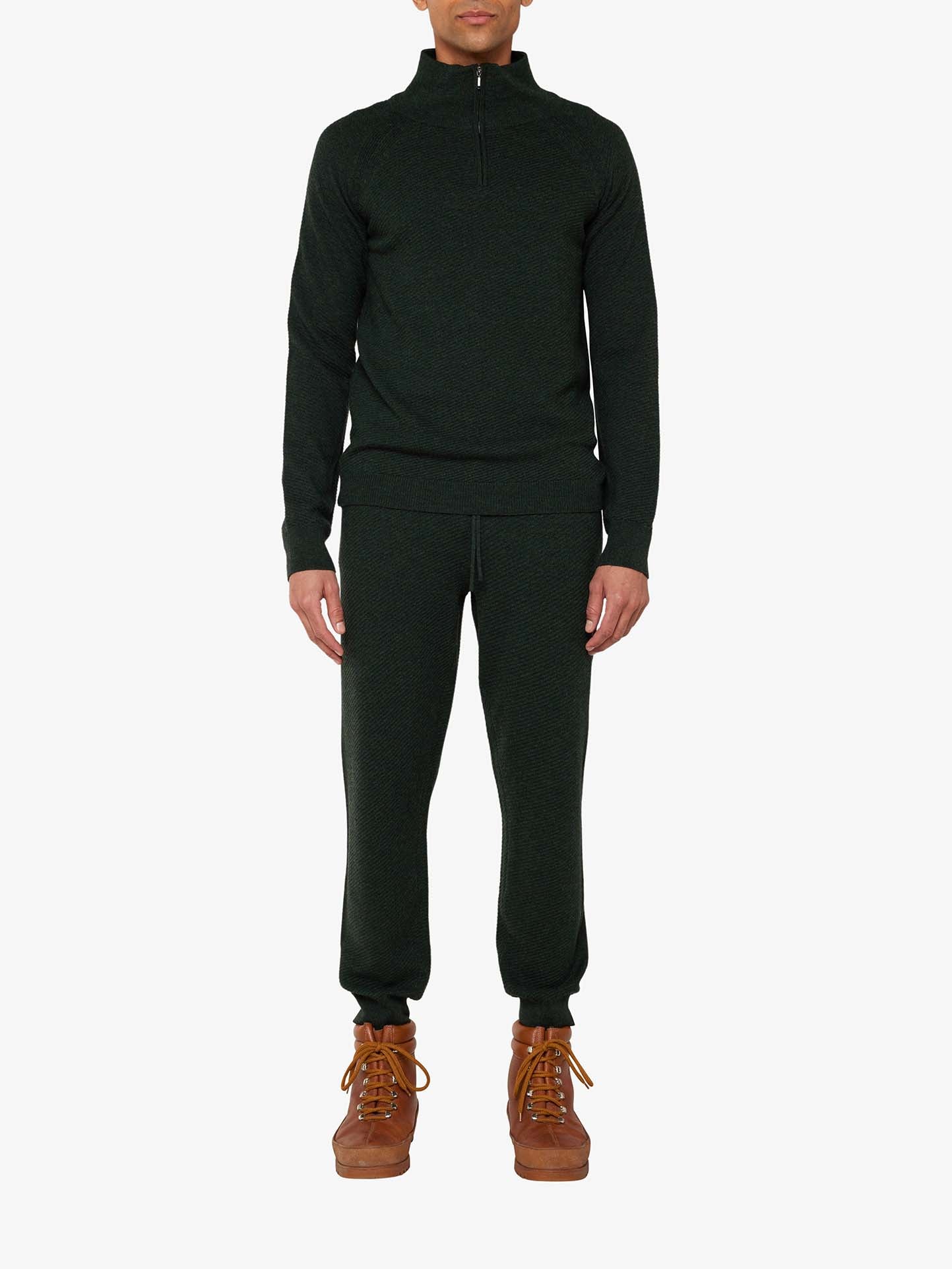A man is standing, showcasing the Geilo Jogger Men by We Norwegians, an elegant lounge set in dark green featuring a half-zip pullover and matching pants. He completes the look with light brown ankle boots against a plain white background.