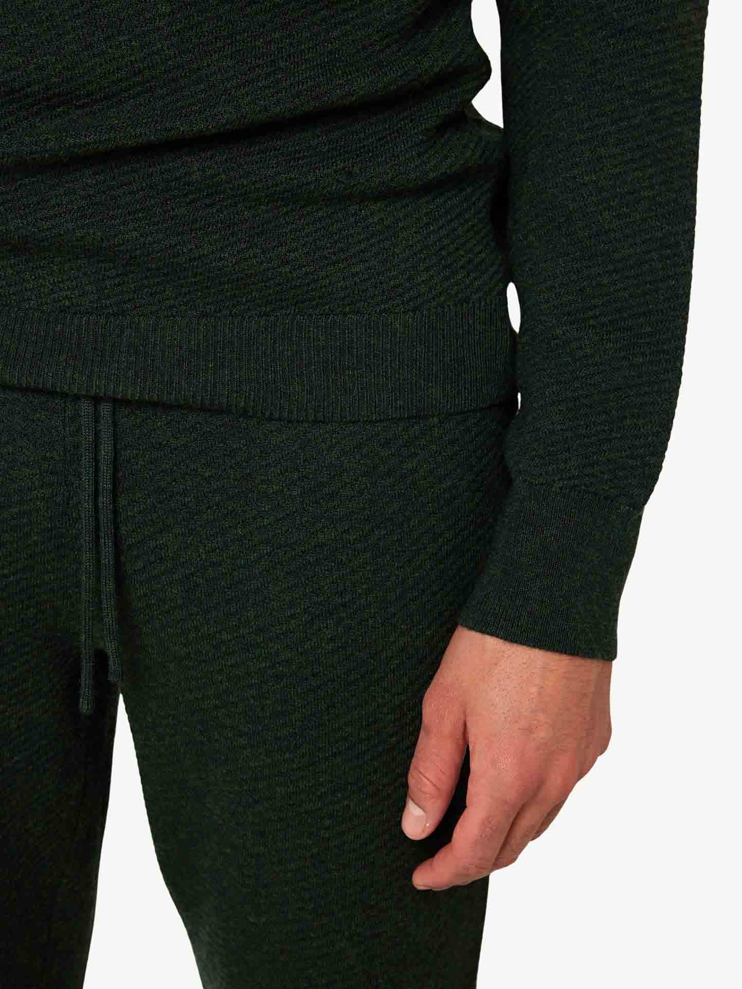 A person is shown from the torso down, wearing a dark green Geilo Jogger Men set by We Norwegians, which includes a fine Merino wool sweater and matching elegant jogger pants. The sleeve of the sweater extends to the wrist, with a hand visible, resting alongside the pants that feature a drawstring waistband.