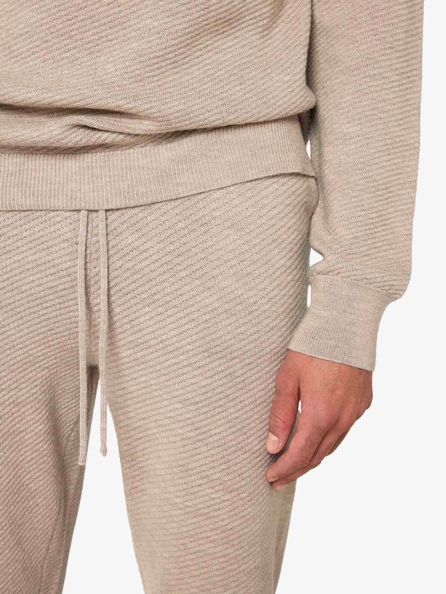 A person is wearing a light beige knit outfit. The image shows a close-up of their torso and legs, highlighting the texture and pattern of the fabric. The matching sweater and We Norwegians Geilo Jogger Men feature ribbed cuffs and an adjustable waist with a drawstring waistband.