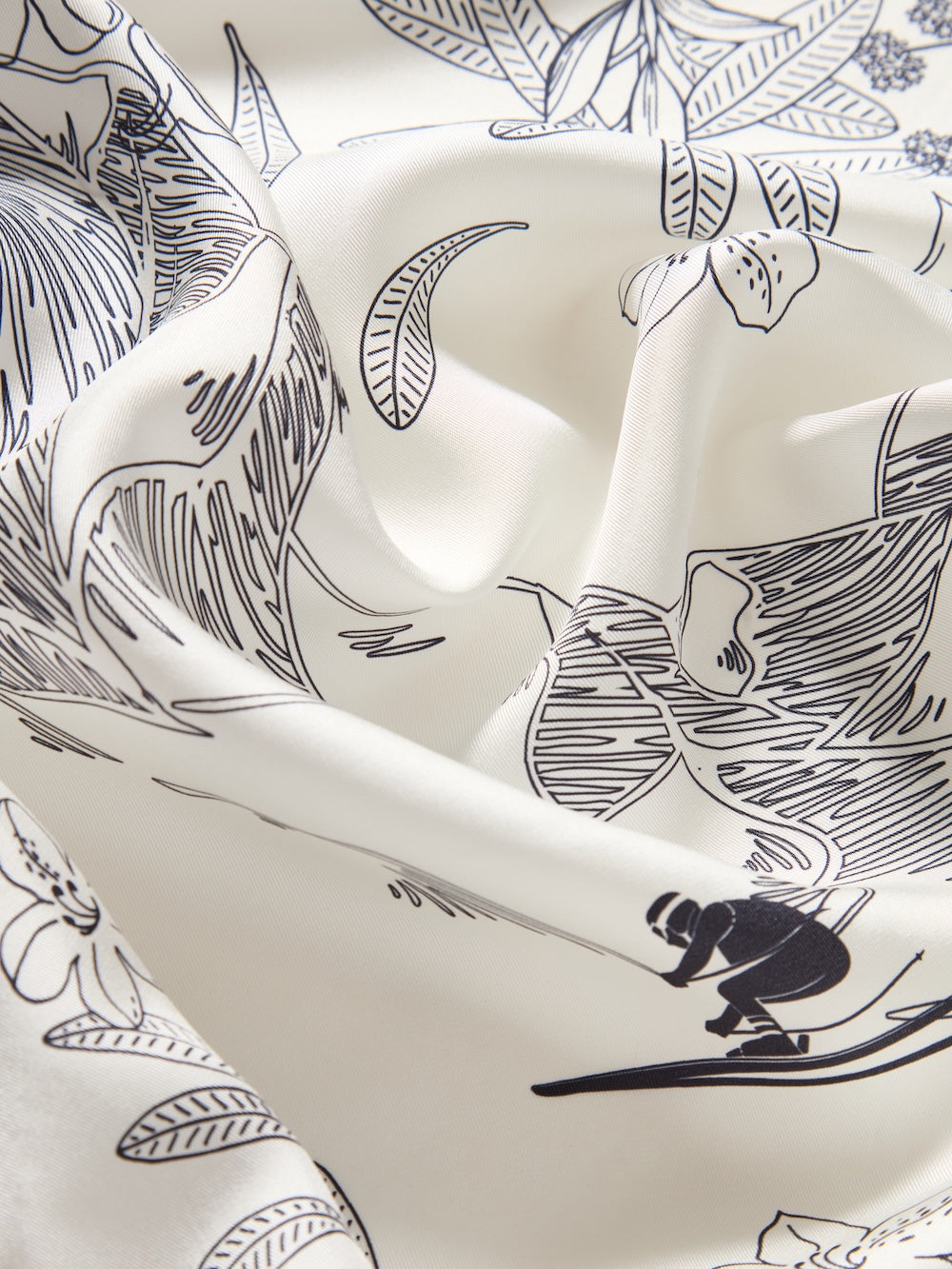 A close-up of the We Norwegians Alps Silk Scarf, showcasing white fabric with black Scandinavian-inspired floral patterns and a skier illustration. The softly folded silk creates gentle shadows and texture.