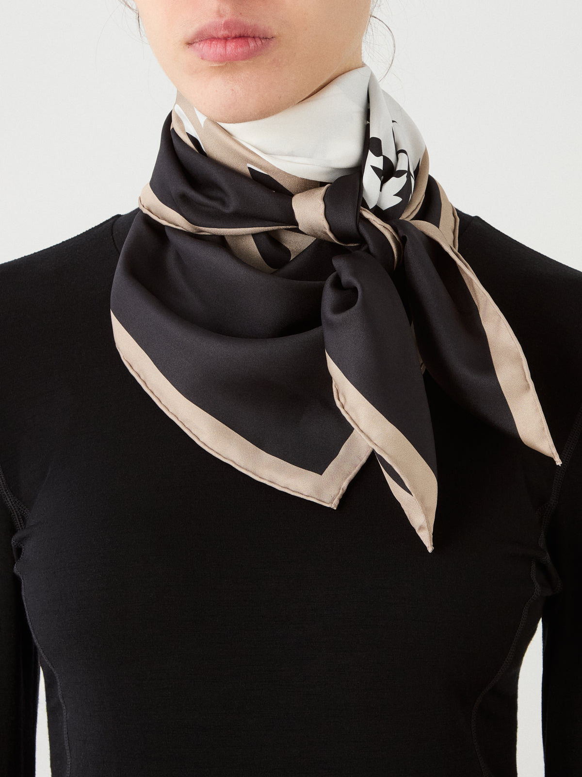 A woman wearing a black top and the We Norwegians Afterski Silk Scarf—featuring a black, beige, and white pattern—neatly tied around her neck. Only the lower half of her face and upper torso are visible.
