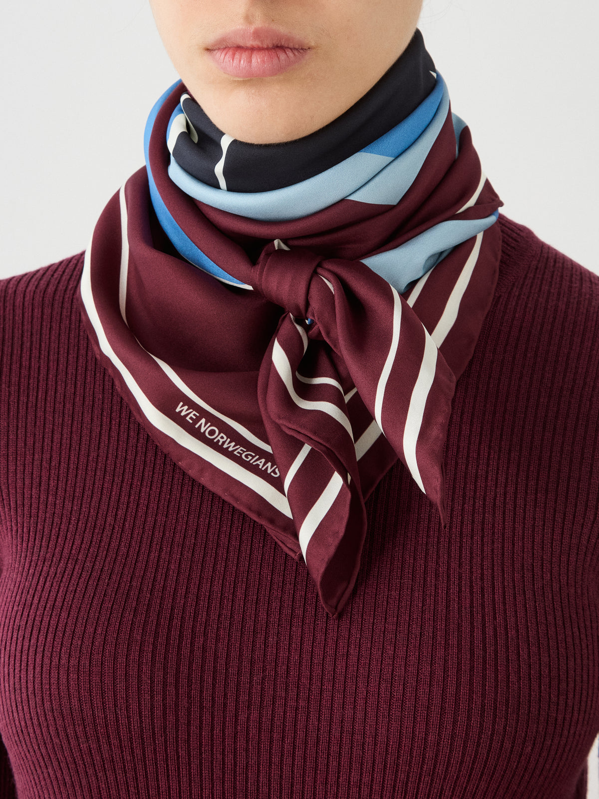 A woman wearing a burgundy ribbed top and the We Norwegians Afterski Silk Scarf—a luxurious silk twill accessory with maroon, white, and blue stripes and “WE NORWEGIANS” printed on one corner—tied around her neck.