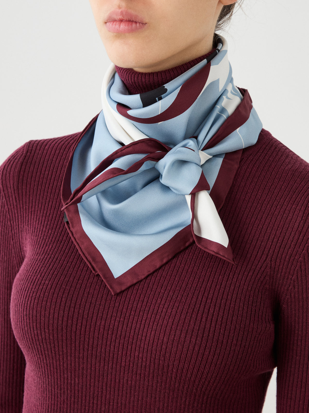 A person wearing a burgundy ribbed turtleneck sweater with the Afterski Silk Scarf by We Norwegians, in blue and burgundy, neatly tied around their neck.