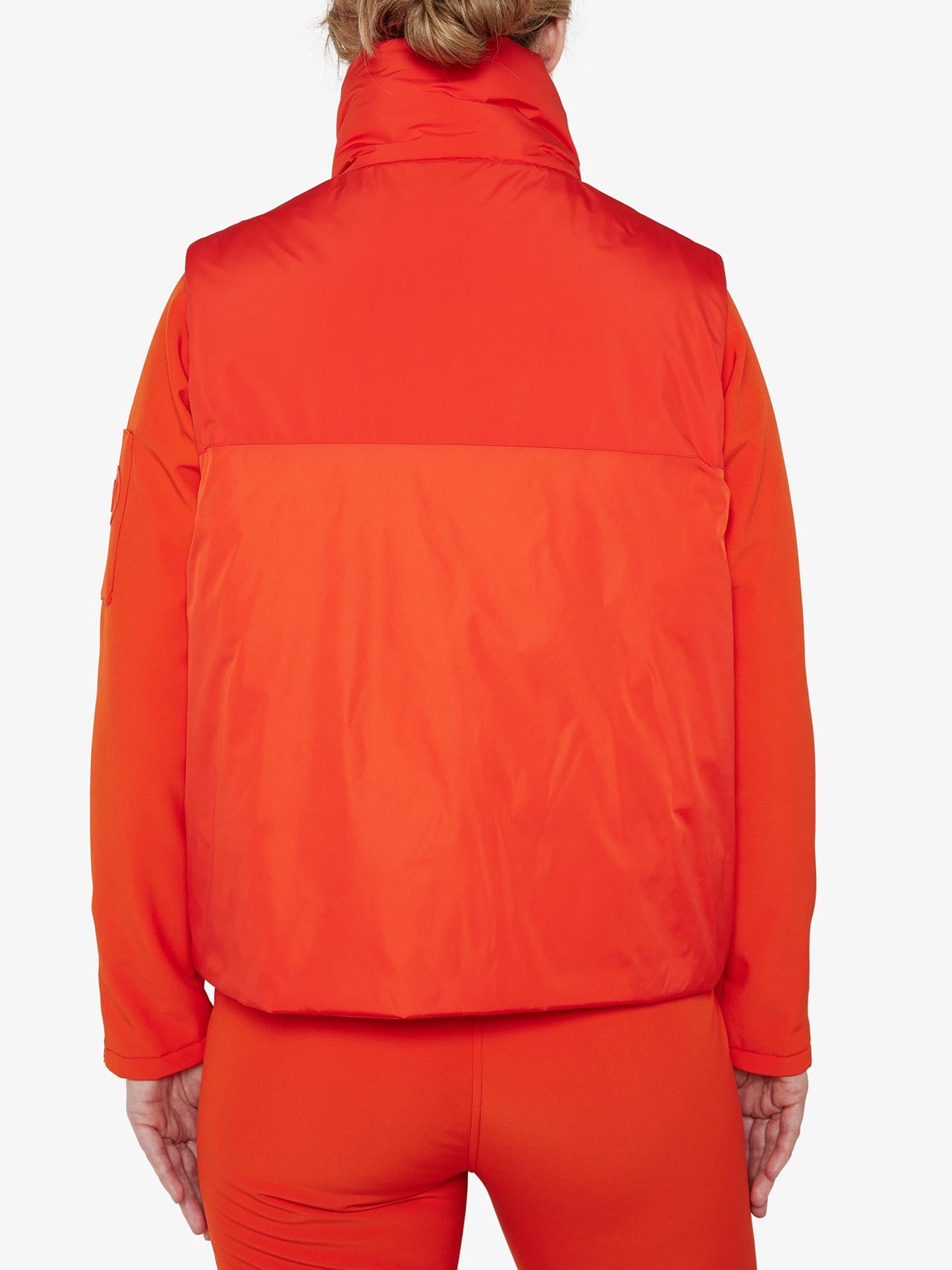 The image displays the back view of a stylish individual dressed in the Filefjell Vest Women by We Norwegians, featuring a vibrant orange jacket paired with matching orange pants. The water-repellent jacket boasts a high collar and a slightly padded appearance. The person's light-colored hair is tied up, set against a white background.