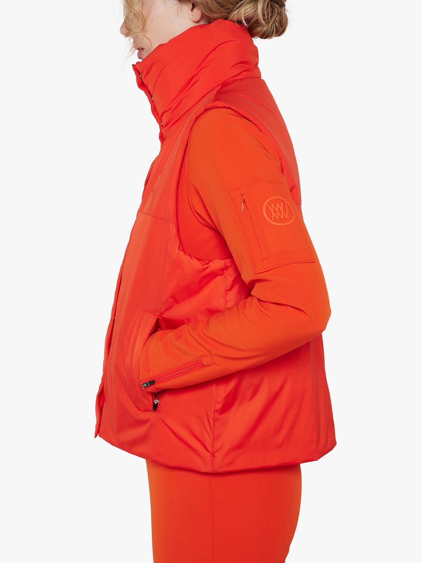 A person with light-colored hair styled in a ponytail is shown from the side wearing a Filefjell Vest Women by We Norwegians. The bright orange puffer vest, made of water-repellent fabric, is worn over an orange long-sleeve shirt and pants. It features a zippered side pocket and a logo patch on the upper arm, against a plain white background.
