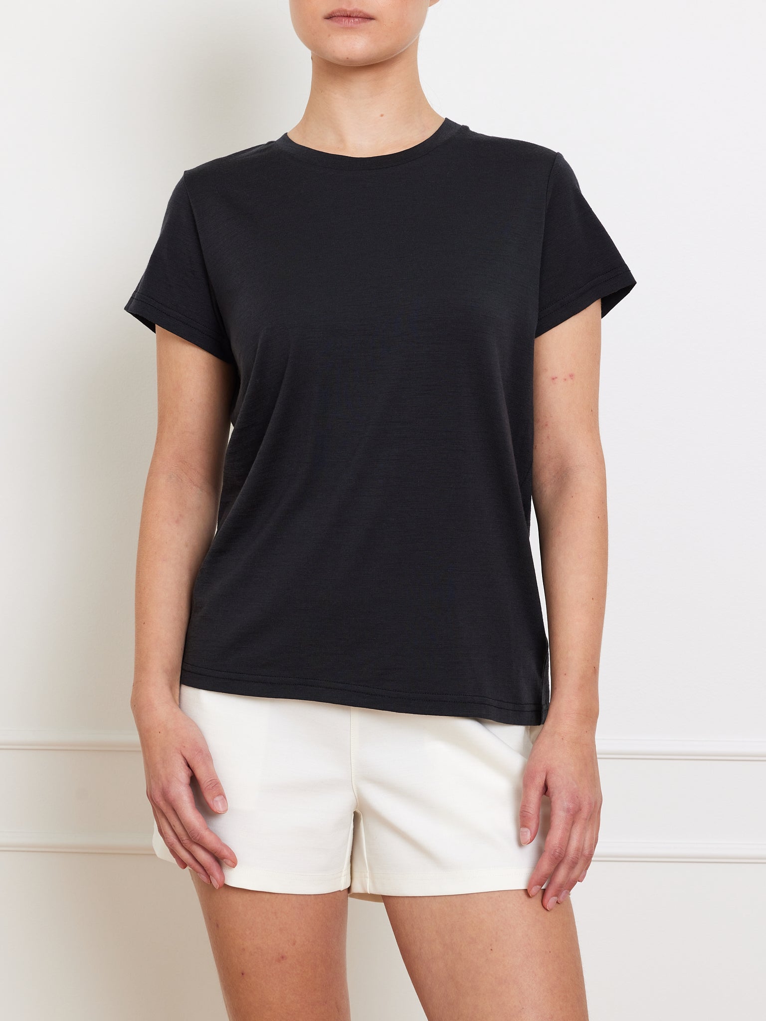 A person wearing a We Norwegians Berg Favorite Tee Women in Merino Tencel and white shorts stands against a light wall, showcasing the no-itch comfort of the shirt. They're shown from shoulders down, highlighting this essential wardrobe piece.