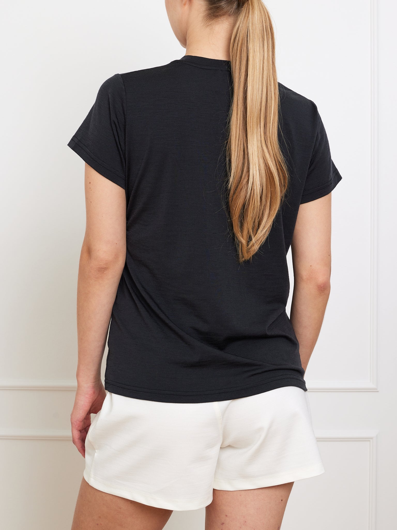Rear view of a person with long blonde hair wearing the black Berg Favorite Tee Women by We Norwegians and white shorts. The no-itch Merino Tencel fabric makes it a wardrobe staple against a plain, light-colored wall.