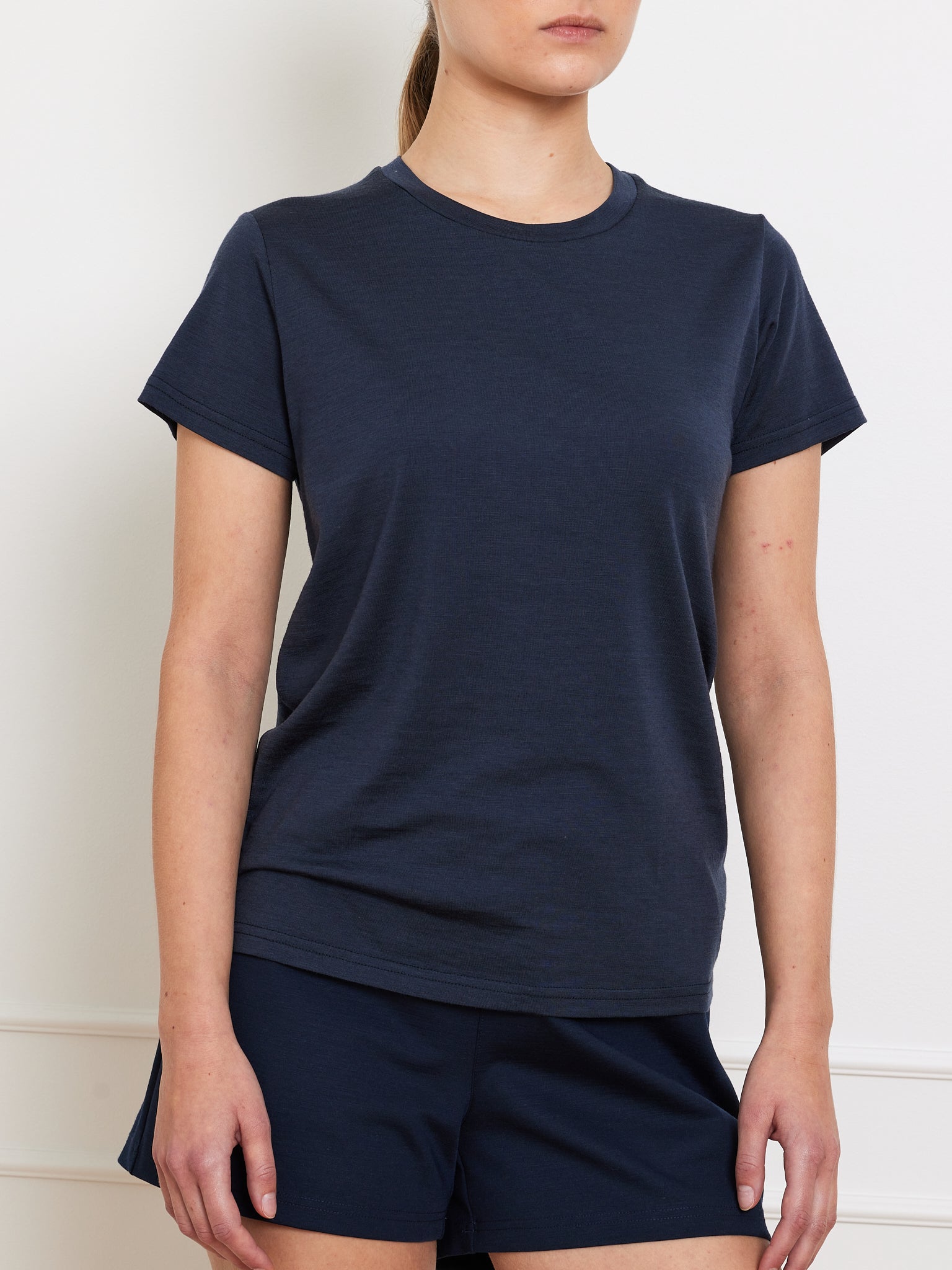 Wearing a dark blue Berg Favorite Tee Women by We Norwegians and matching shorts, someone stands against a plain white wall. Their arms hang by their sides, partially obscuring their face. This no-itch fabric outfit is a casual wardrobe staple.