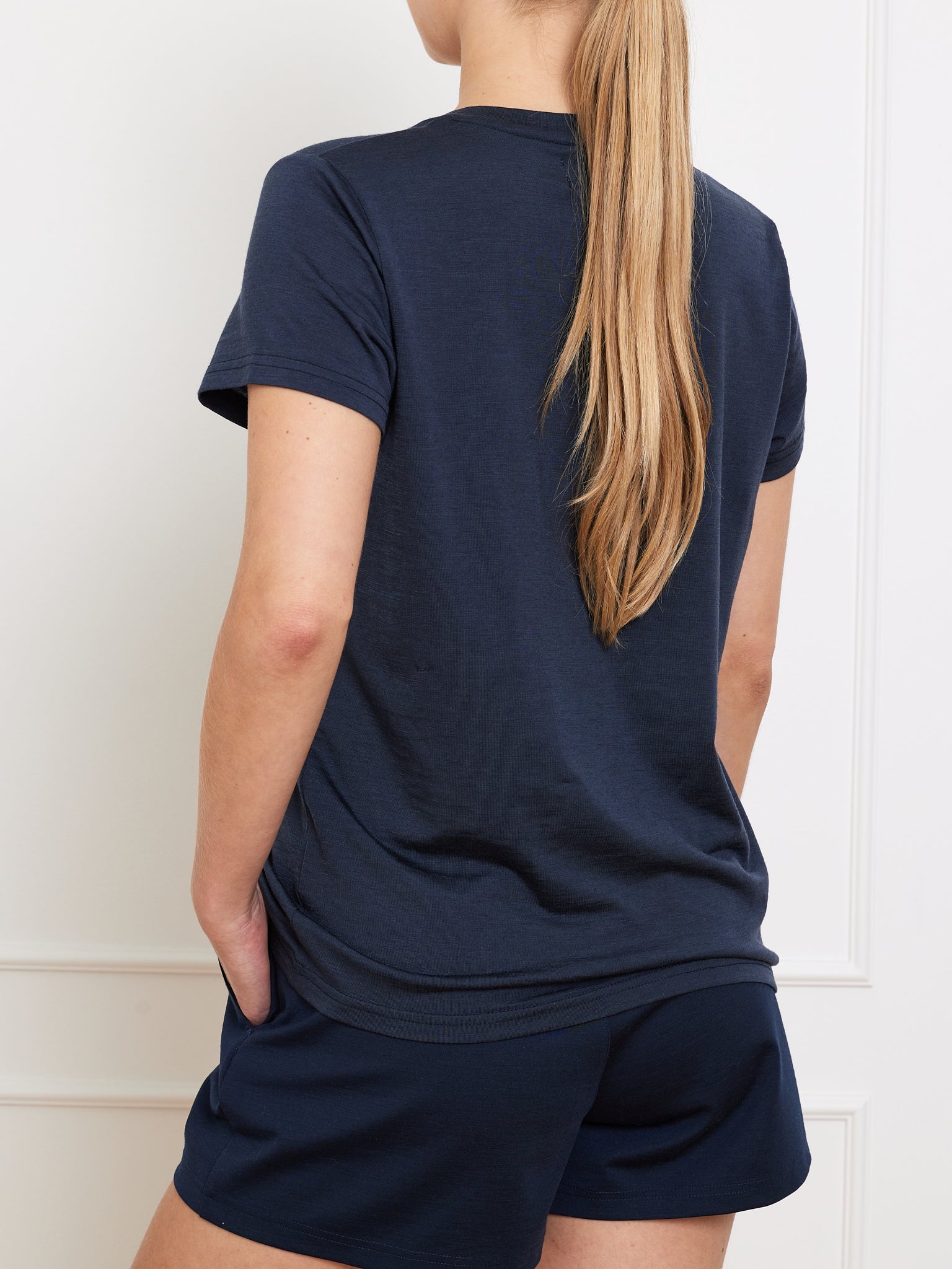 A person with long, straight hair showcases the Berg Favorite Tee Women by We Norwegians and shorts in navy blue, essential wardrobe pieces. Their hands rest in the pockets as they casually lean against a white wall.