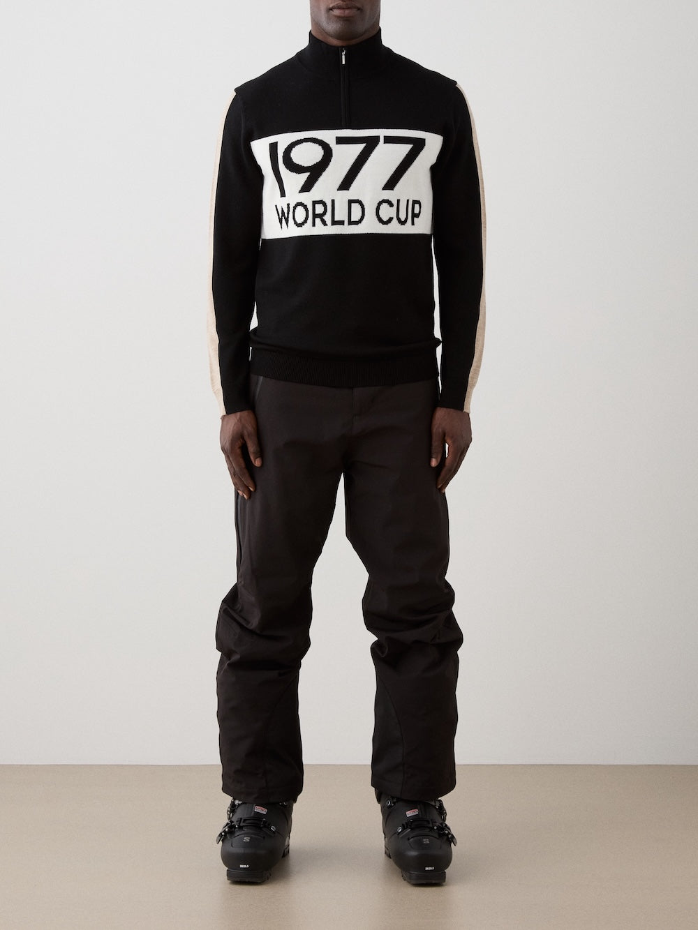 A person stands indoors in black ski pants, ski boots, and a We Norwegians 1977 World Cup Zipup Men sweater with "1977 WORLD CUP" boldly on the front. The minimal background highlights this classic alpine style.