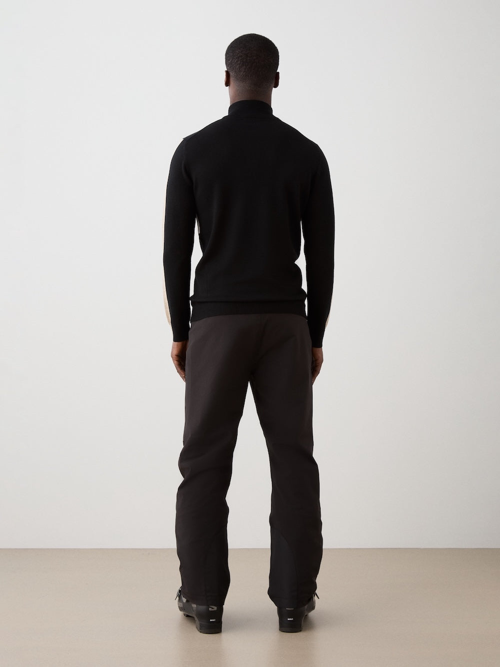 A man faces away from the camera, wearing the We Norwegians 1977 World Cup Zipup Men sweater in black with dark pants, standing against a plain light-colored wall and floor.
