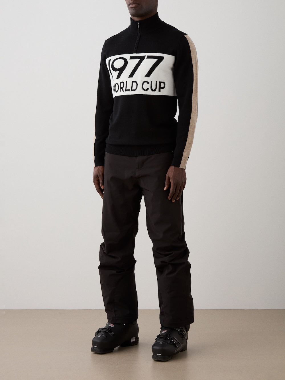 A person wearing the We Norwegians 1977 World Cup Zipup Men in black and white, along with black pants and black ski boots, stands indoors against a plain background.