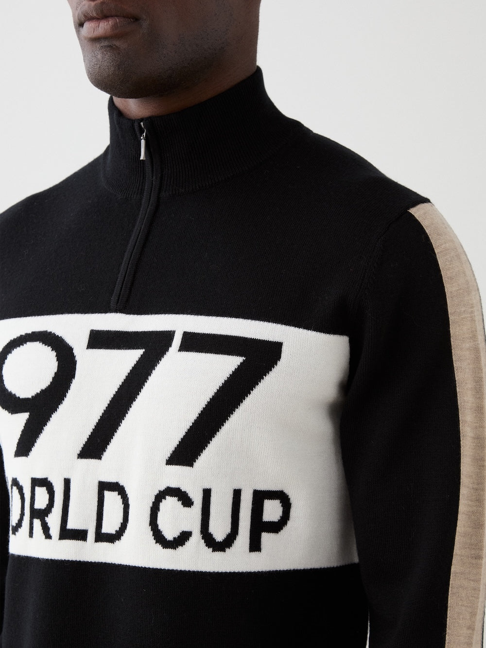 A man wears the We Norwegians 1977 World Cup Zipup Men ski sweater, featuring a black alpine style with beige sleeve accents and a bold "1977 WORLD CUP" white chest panel.