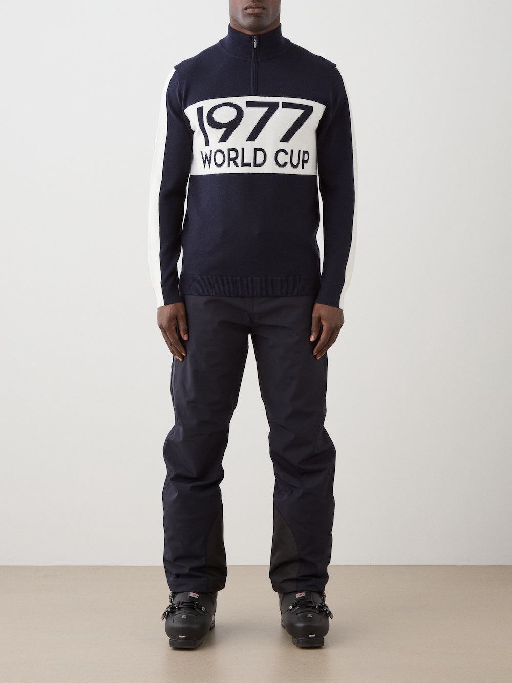 A person stands against a plain background, wearing the We Norwegians 1977 World Cup Zipup Men sweater with "1977 WORLD CUP" in bold white letters, paired with dark pants and black ski boots for a classic alpine-inspired look.