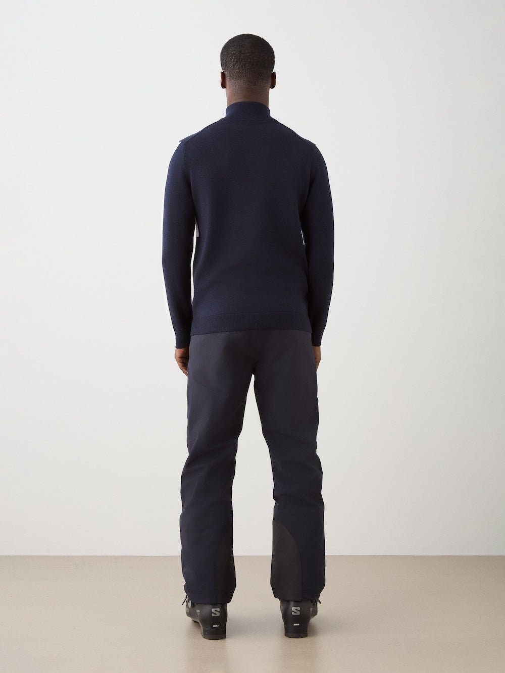 A man stands indoors facing away from the camera, wearing a We Norwegians 1977 World Cup Zipup Men sweater with dark pants and black boots, set against a plain light background.
