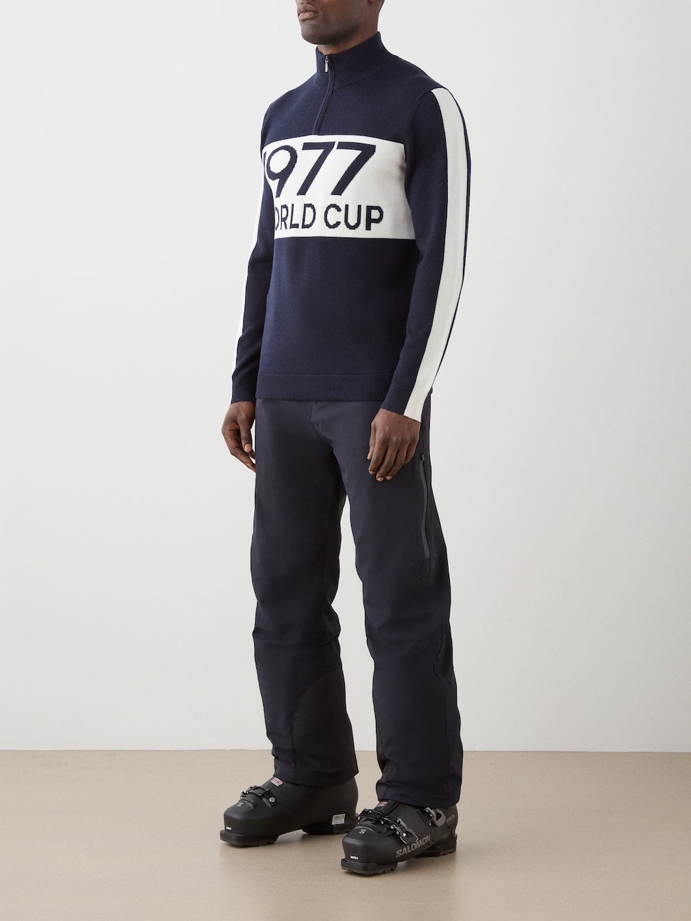 A man stands indoors wearing We Norwegians’ 1977 World Cup Zipup Men in navy blue and white, styled with navy pants and black ski boots.
