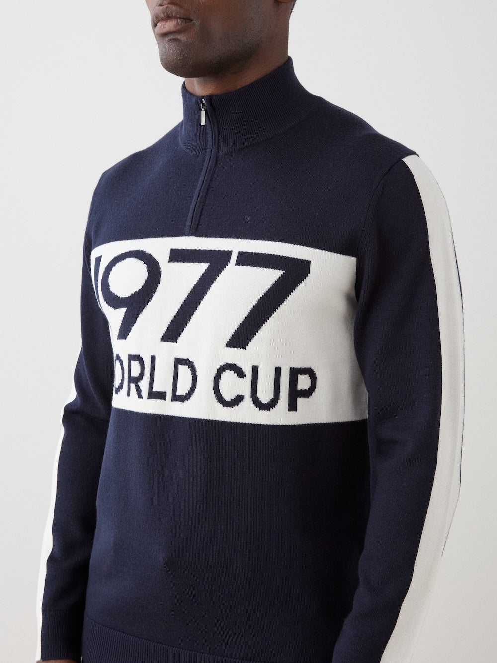 A person wears the We Norwegians 1977 World Cup Zipup Men—a navy alpine ski sweater with a white chest panel reading "1977 WORLD CUP," quarter-zip, and white sleeve stripes.