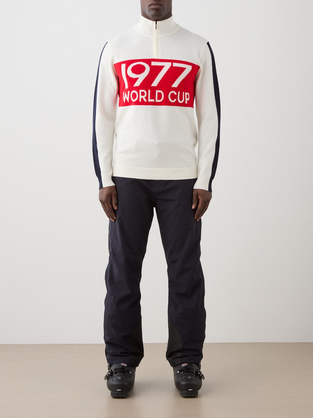 A person wearing the We Norwegians 1977 World Cup Zipup Men with a red "1977 WORLD CUP" graphic, dark striped pants, and black ski boots stands against a plain background.