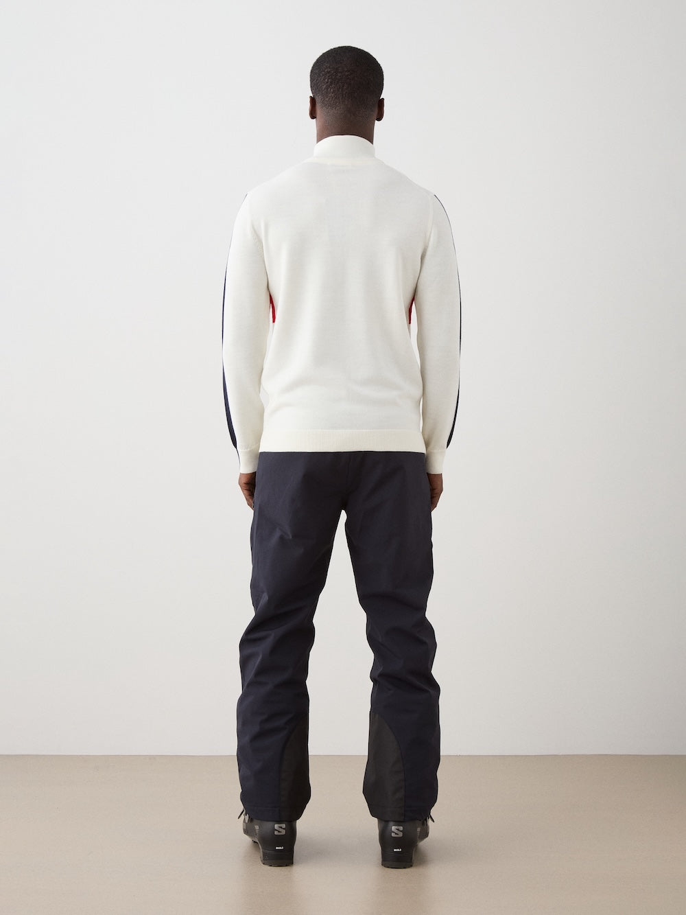 A man faces away from the camera, wearing the We Norwegians 1977 World Cup Zipup Men sweater with dark pants and black shoes, set against a plain light-colored wall and floor.