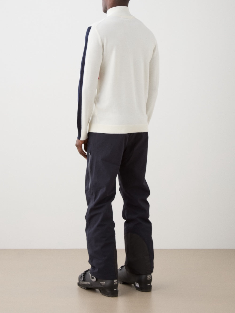 A person stands facing away, wearing the We Norwegians 1977 World Cup Zipup Men ski sweater, dark snow pants, and black ski boots, against a plain light background.