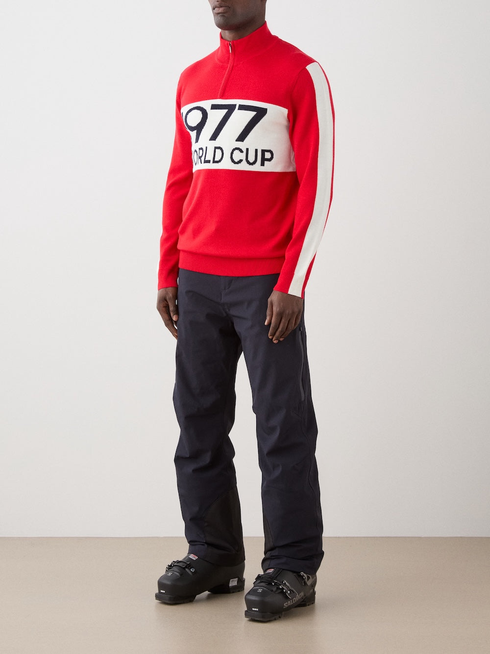 A man wears the We Norwegians 1977 World Cup Zipup Men in red and white, featuring "1977 WORLD CUP" on the front, paired with black pants and black snow boots for a stylish alpine ski look against a plain background.