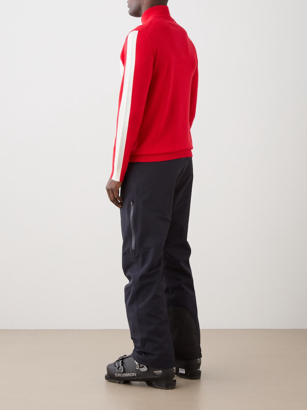 A person stands facing away, wearing the We Norwegians 1977 World Cup Zipup Men sweater with white sleeve stripes, black pants, and black Salomon ski boots against a plain light background.