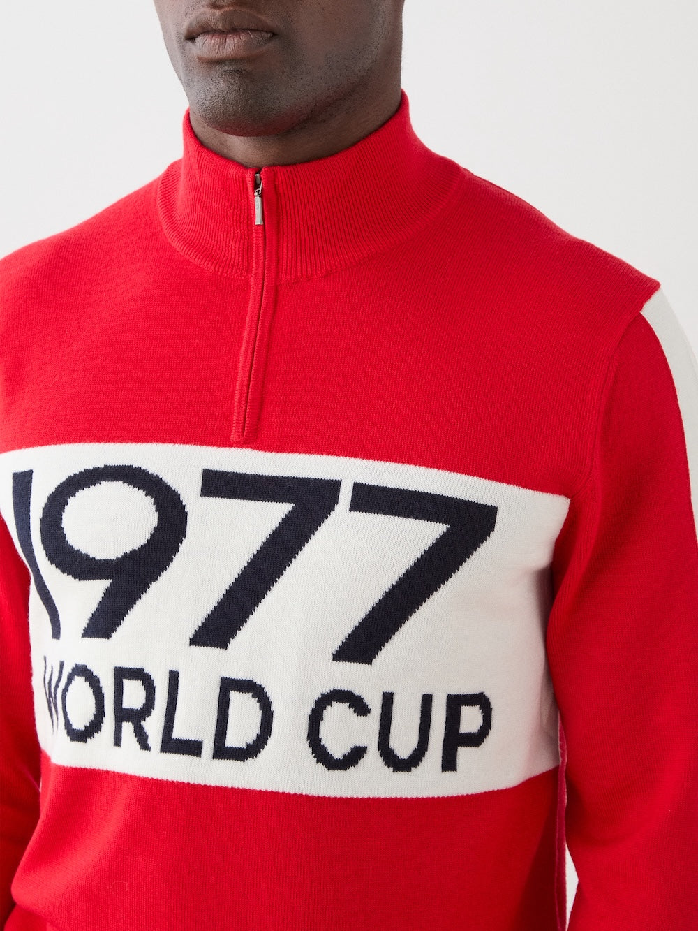 A person wears the We Norwegians 1977 World Cup Zipup Men—an alpine-style red ski sweater with a white chest panel displaying "1977 WORLD CUP" in bold black text; only their lower face and upper torso are visible.