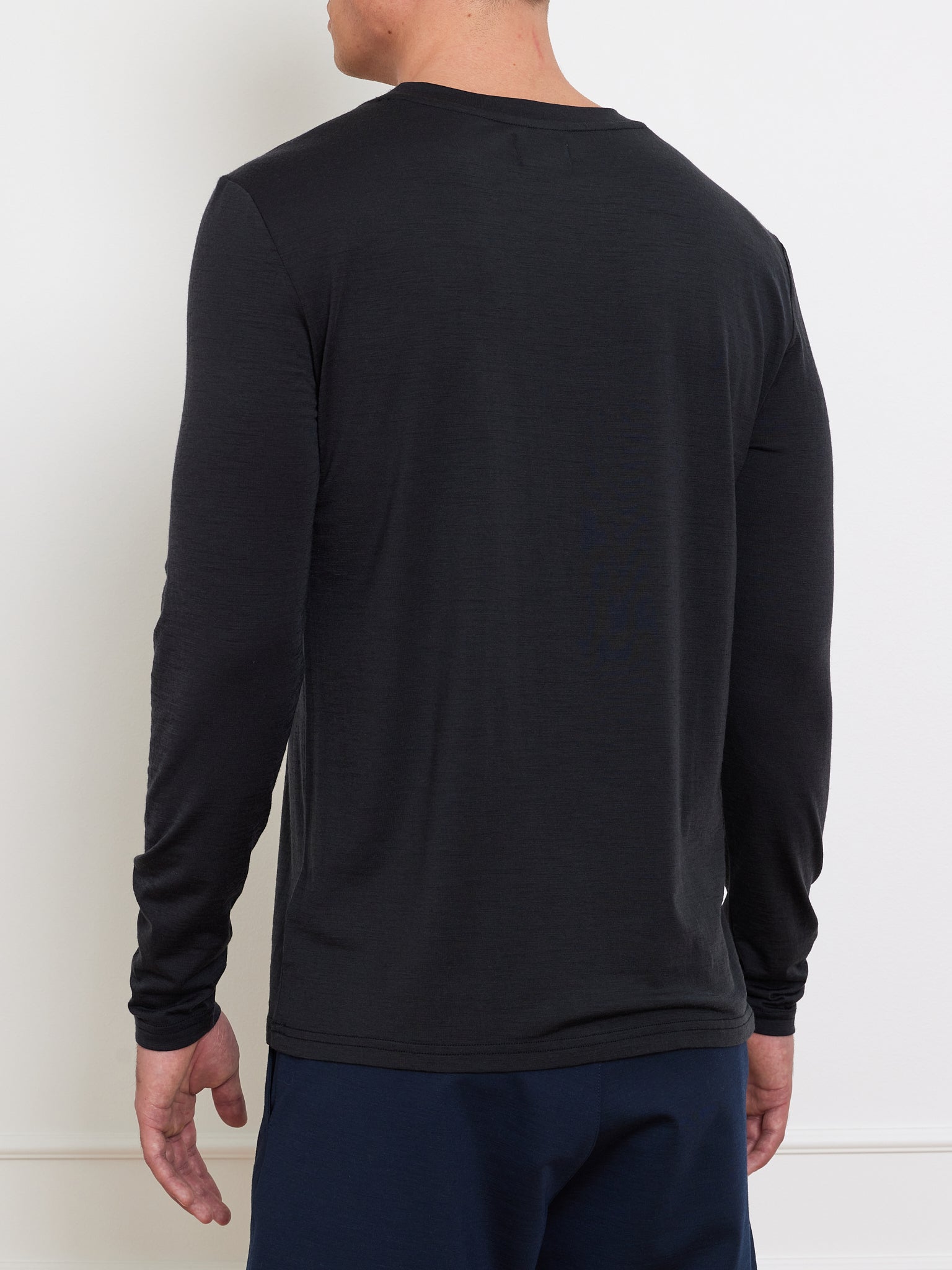 A person stands with their back to the camera, wearing a We Norwegians Berg LongSleeve Men in plain black and dark blue pants against a stark white background.
