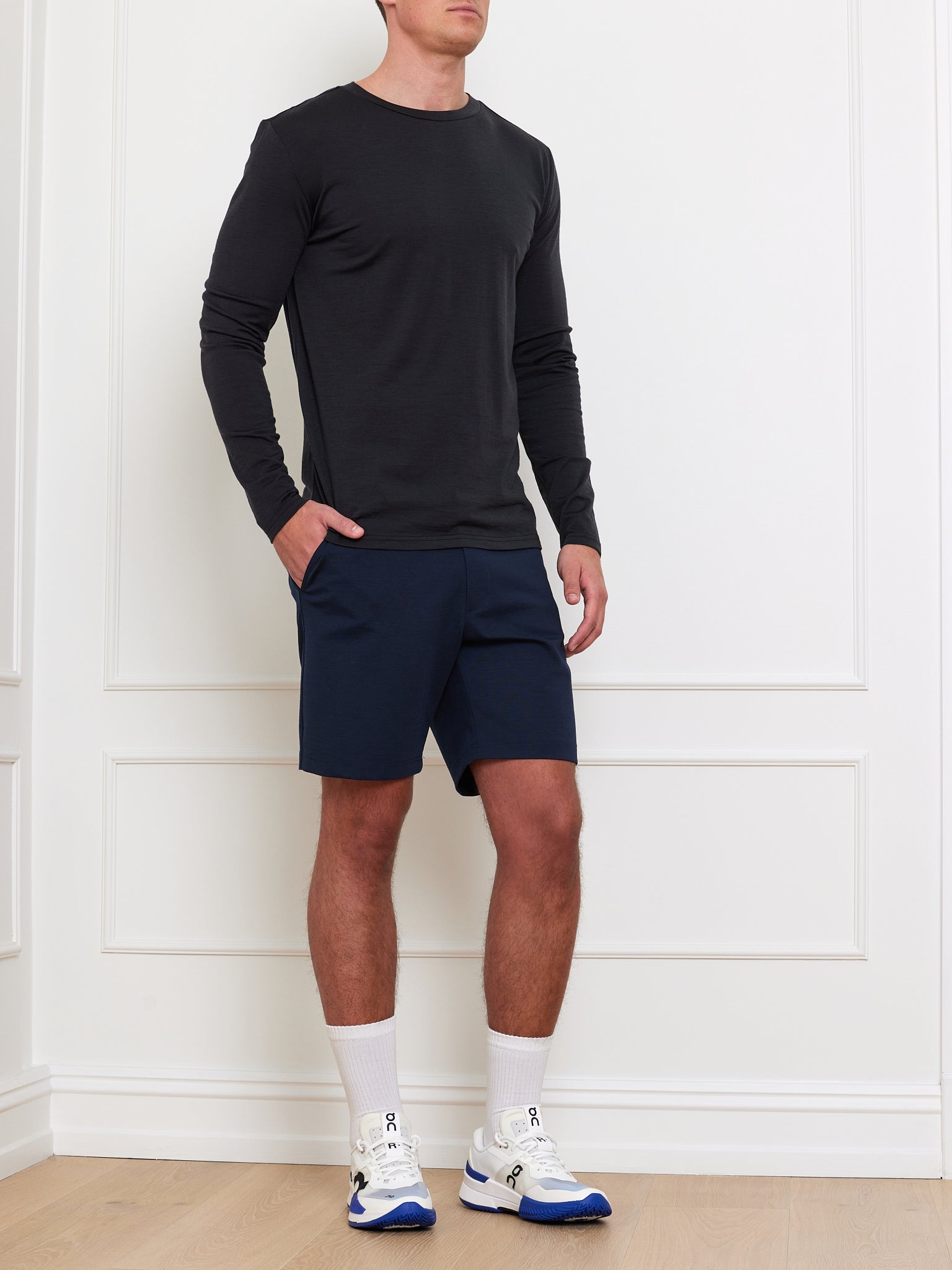 A person in a black Berg LongSleeve Men shirt by We Norwegians, navy shorts, and white sneakers with blue accents stands against a white wall. Hands in pockets, they face forward, highlighting a casual look ideal for layering with merino wool base layers.