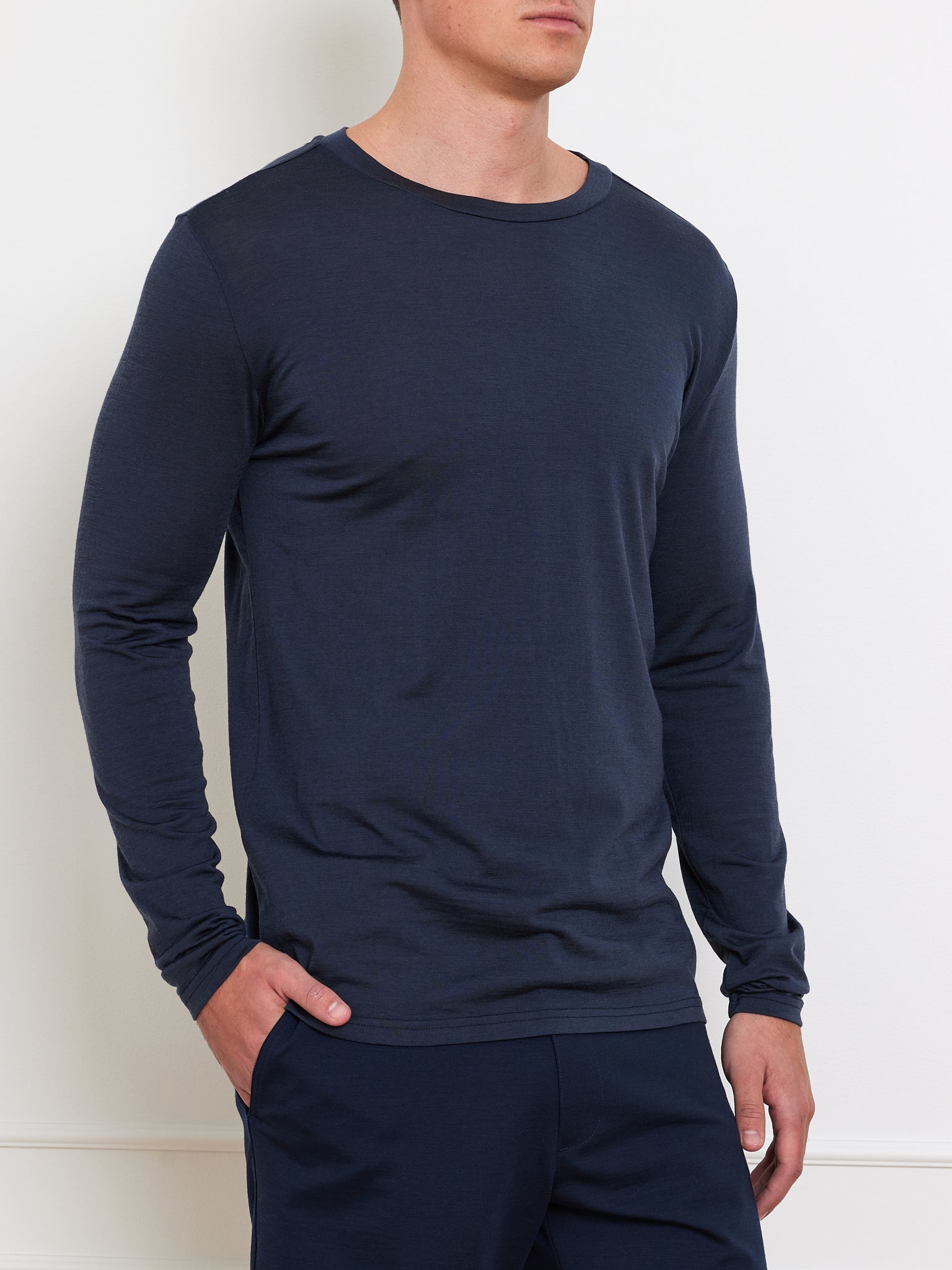 A person models the We Norwegians Berg LongSleeve Men in dark blue, a soft merino wool ensemble including matching pants. Set against a white backdrop, they stand with their left hand in their pocket. The close-fitting shirt highlights the smooth base layer as they slightly turn to the left.