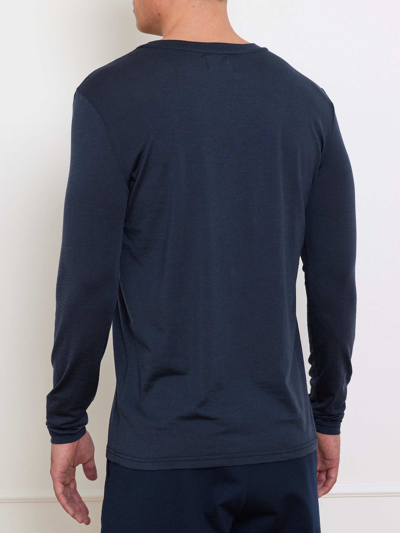 A person in a dark blue We Norwegians Berg LongSleeve shirt and matching pants is standing against a plain white wall, facing away from the camera.