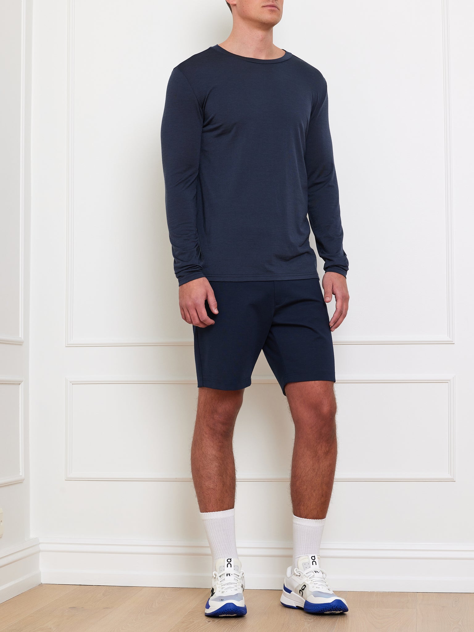 A person stands against a white wall, wearing the navy Berg LongSleeve Men shirt by We Norwegians, paired with matching shorts. They sport white socks and sneakers with blue accents. The wooden floor adds warmth to the scene.
