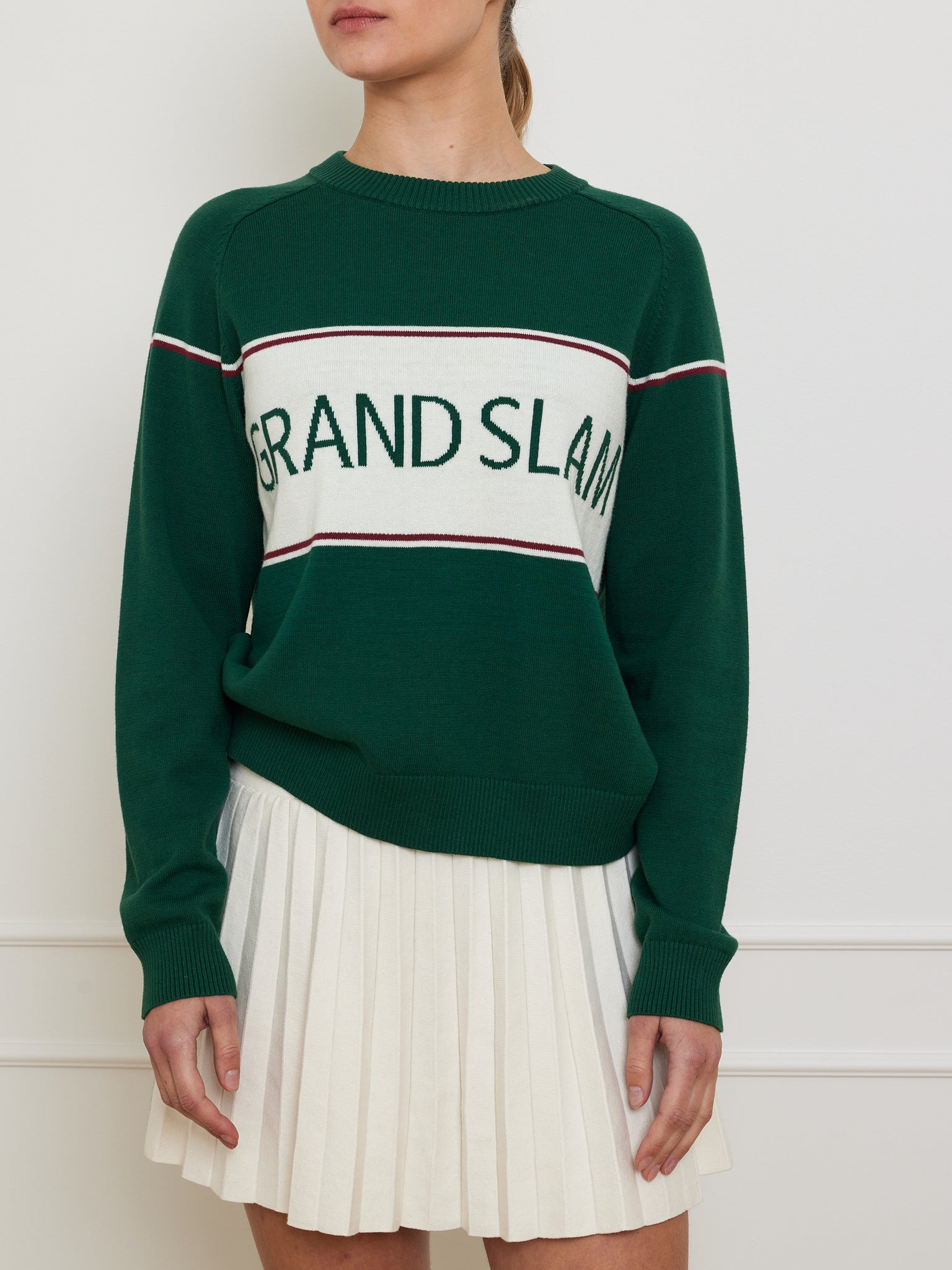 A person wears a We Norwegians Grand Slam Sweater in a vintage athletic style, featuring green with a white "GRAND SLAM" panel and red-white stripes, paired with a white pleated skirt against a plain wall.
