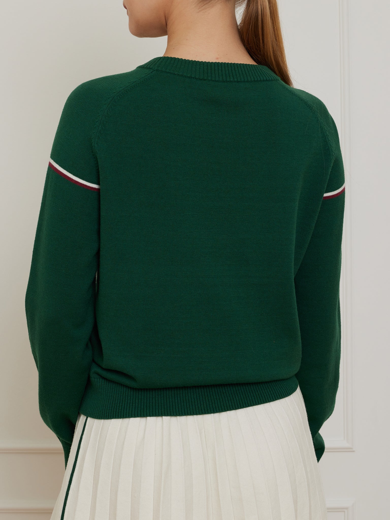 Dressed in a We Norwegians Grand Slam Sweater Women in dark green with white and red striped sleeves, teamed with a cream pleated skirt, the individual showcases a vintage athletic vibe. From behind, their hair is in a ponytail set against a light-colored wall.