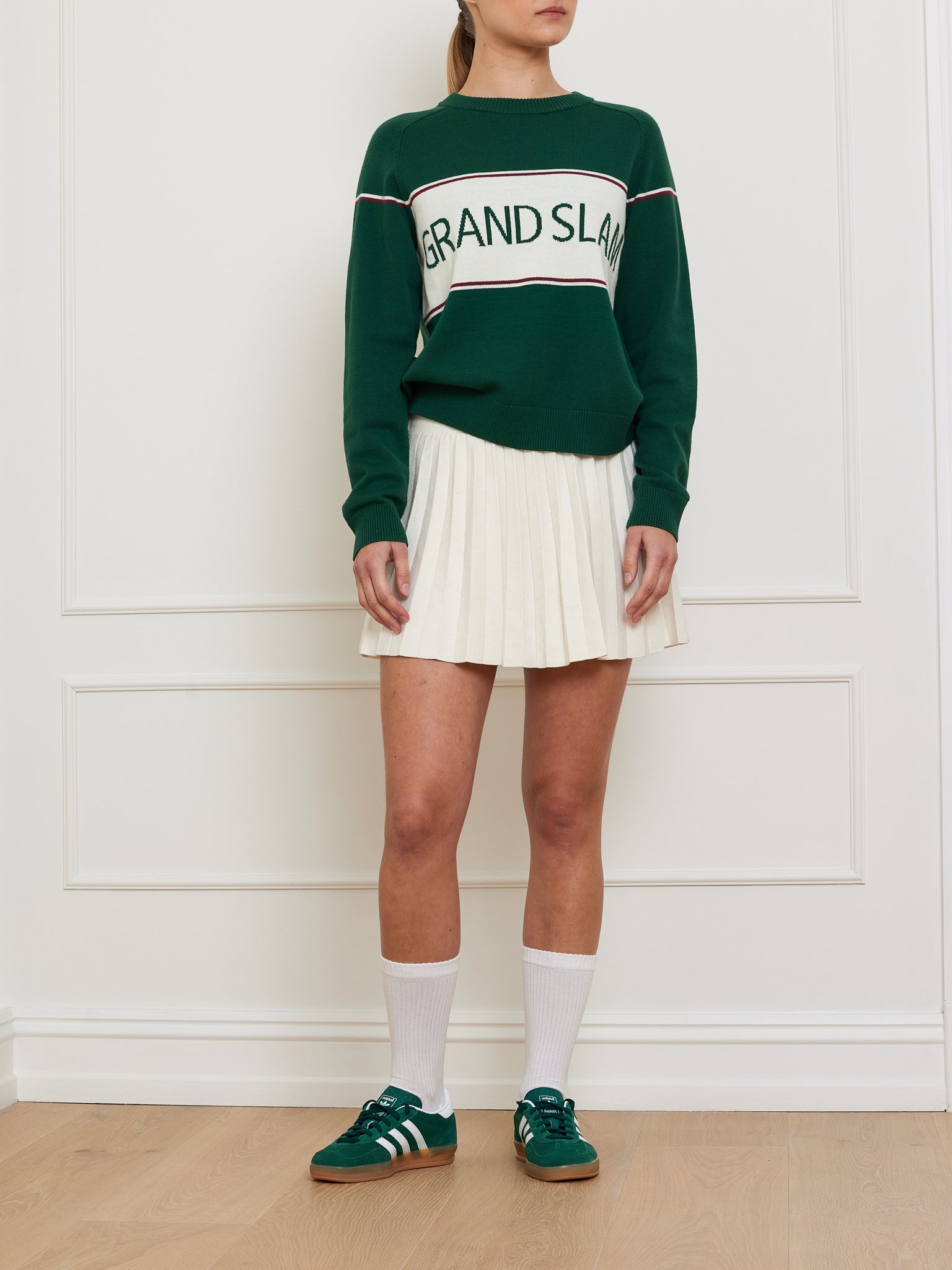 Against a white wall, a person wears a We Norwegians "Grand Slam Sweater Women" made from a cotton-merino blend. With a white pleated skirt, knee-high socks, and green sneakers with white stripes, the look achieves an effortlessly classic sporty vintage athletic style.