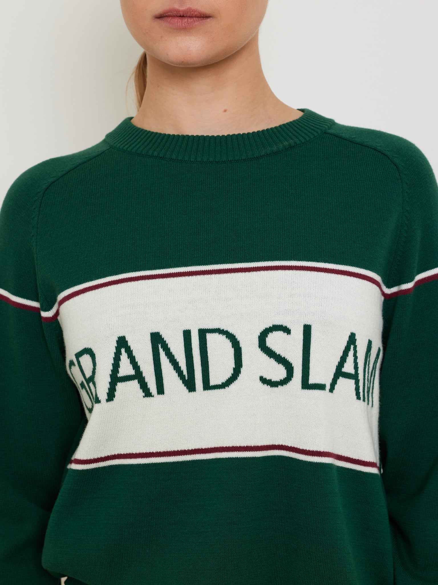 A person sports a We Norwegians Grand Slam Sweater Women, a vintage athletic style in a cotton-merino blend. The green sweater has a white panel with "GRAND SLAM" in green letters and thin red stripes, set against a plain background.