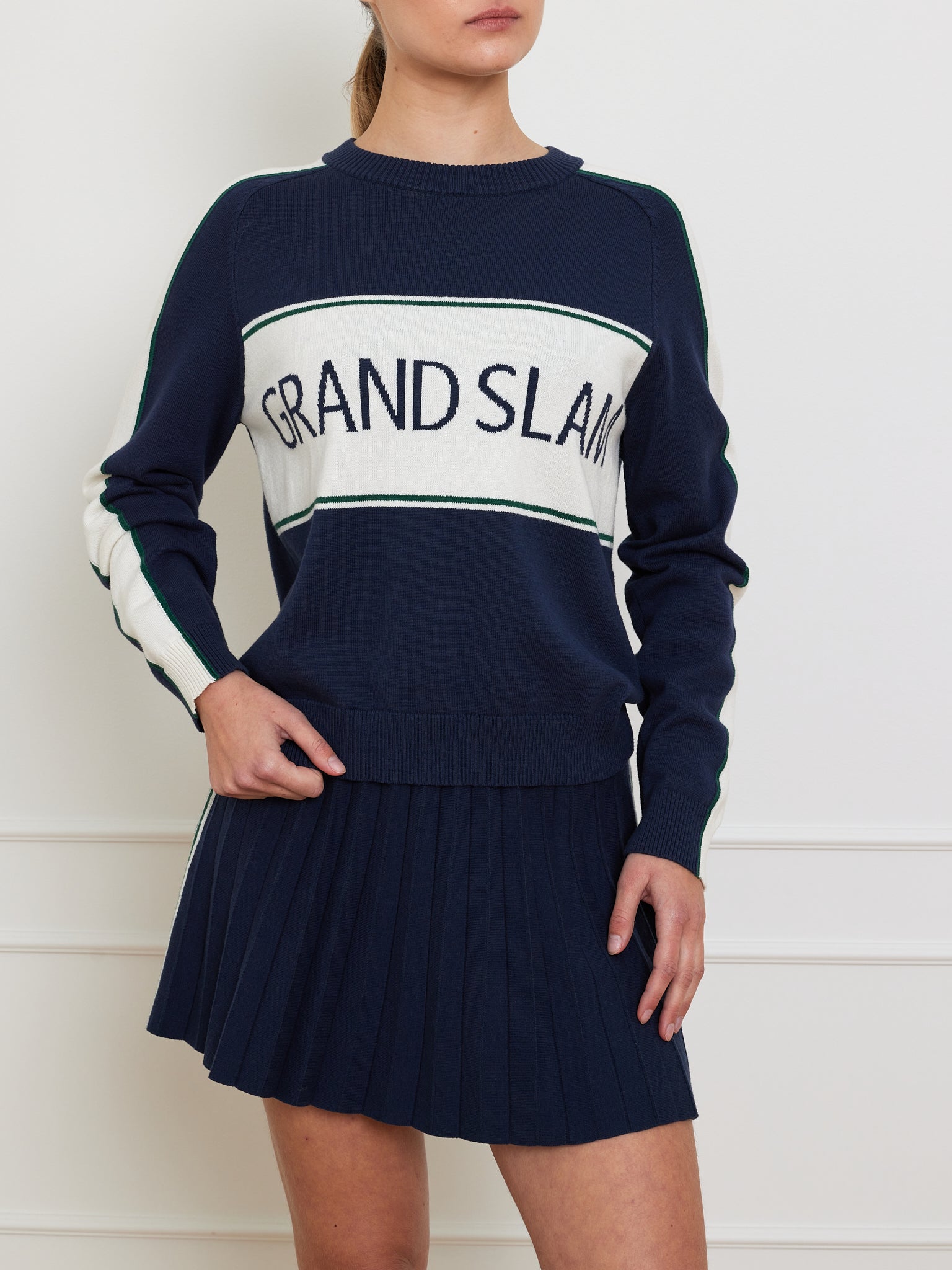 A person wears a Grand Slam Sweater Women by We Norwegians in rich navy cotton-merino with white sleeve and chest accents, paired with a matching pleated skirt. The vintage athletic style shines as they stand against a white wall.