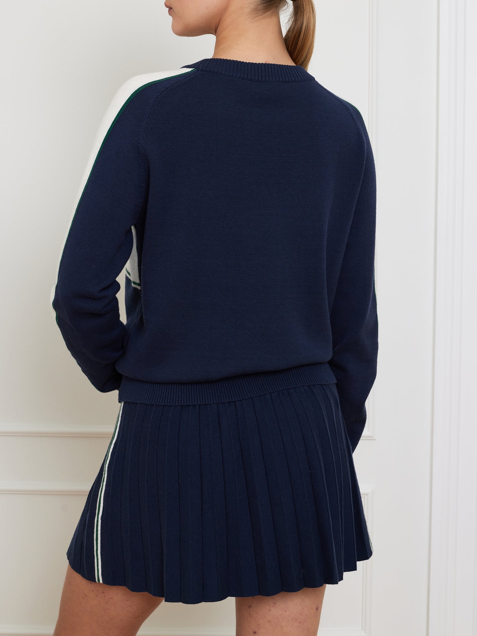 A person wearing a navy We Norwegians Grand Slam Sweater Women with white accents and a matching navy pleated skirt stands facing away, set against a white paneled wall. The vintage athletic style outfit highlights its classic charm in every cotton-merino blend stitch.