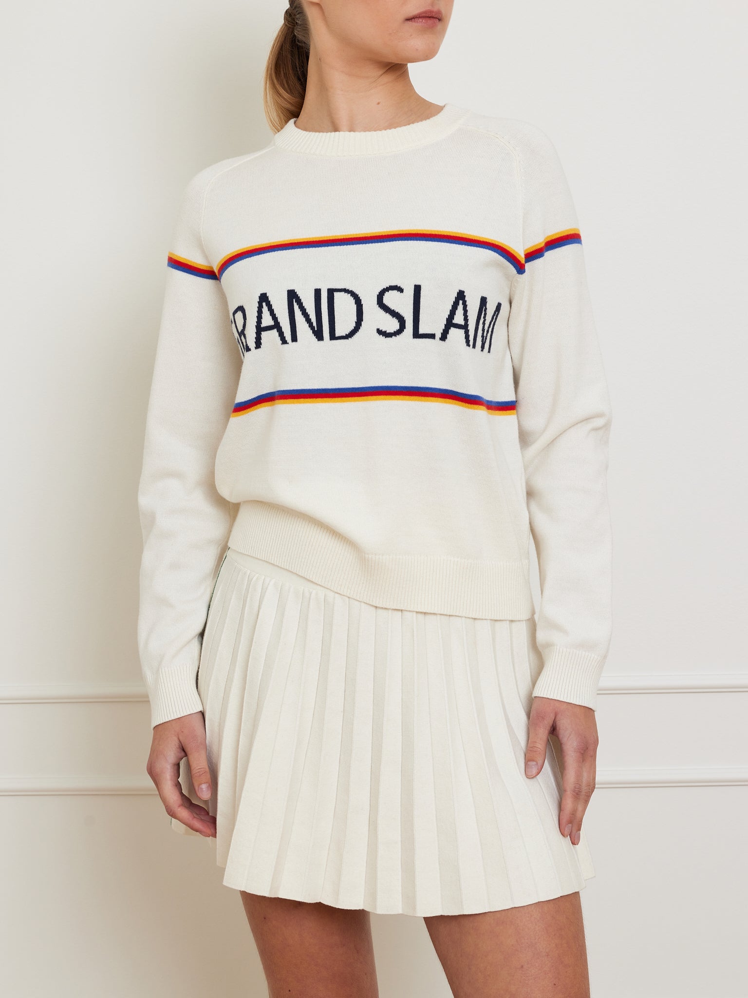 A wearer dons the vintage-style Grand Slam Sweater by We Norwegians, made from a cozy cotton-merino blend. The white long-sleeve design features bold "GRAND SLAM" lettering and is adorned with blue, red, and yellow stripes. They complete the look with a white pleated skirt.